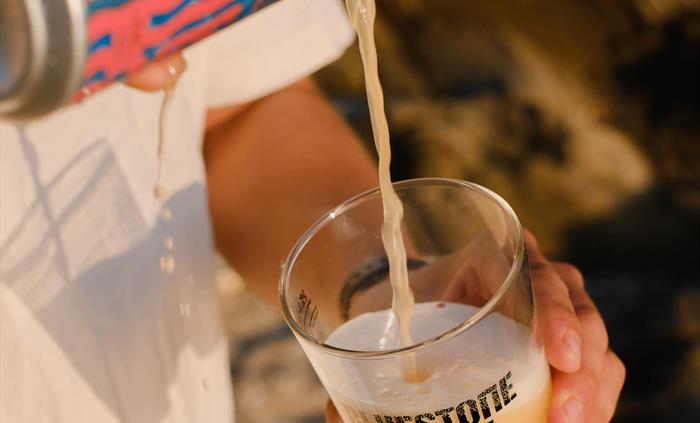 A can of beer is being poured into a glass on a sunny day, the glass has the Bluestone Brewing Co logo printed on the side